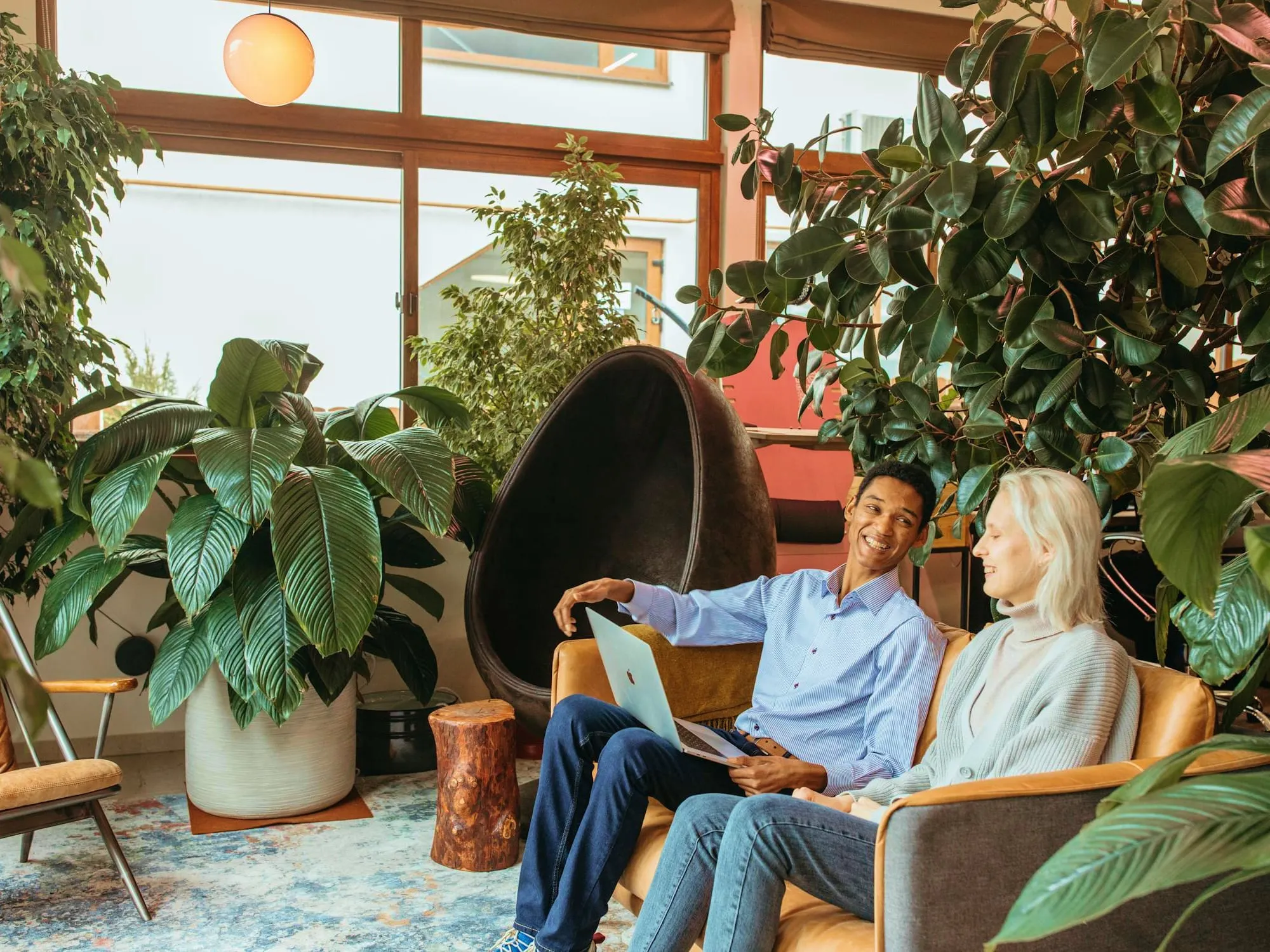 Two people sitting on a sofa surrounded by plants, chatting with an open laptop.